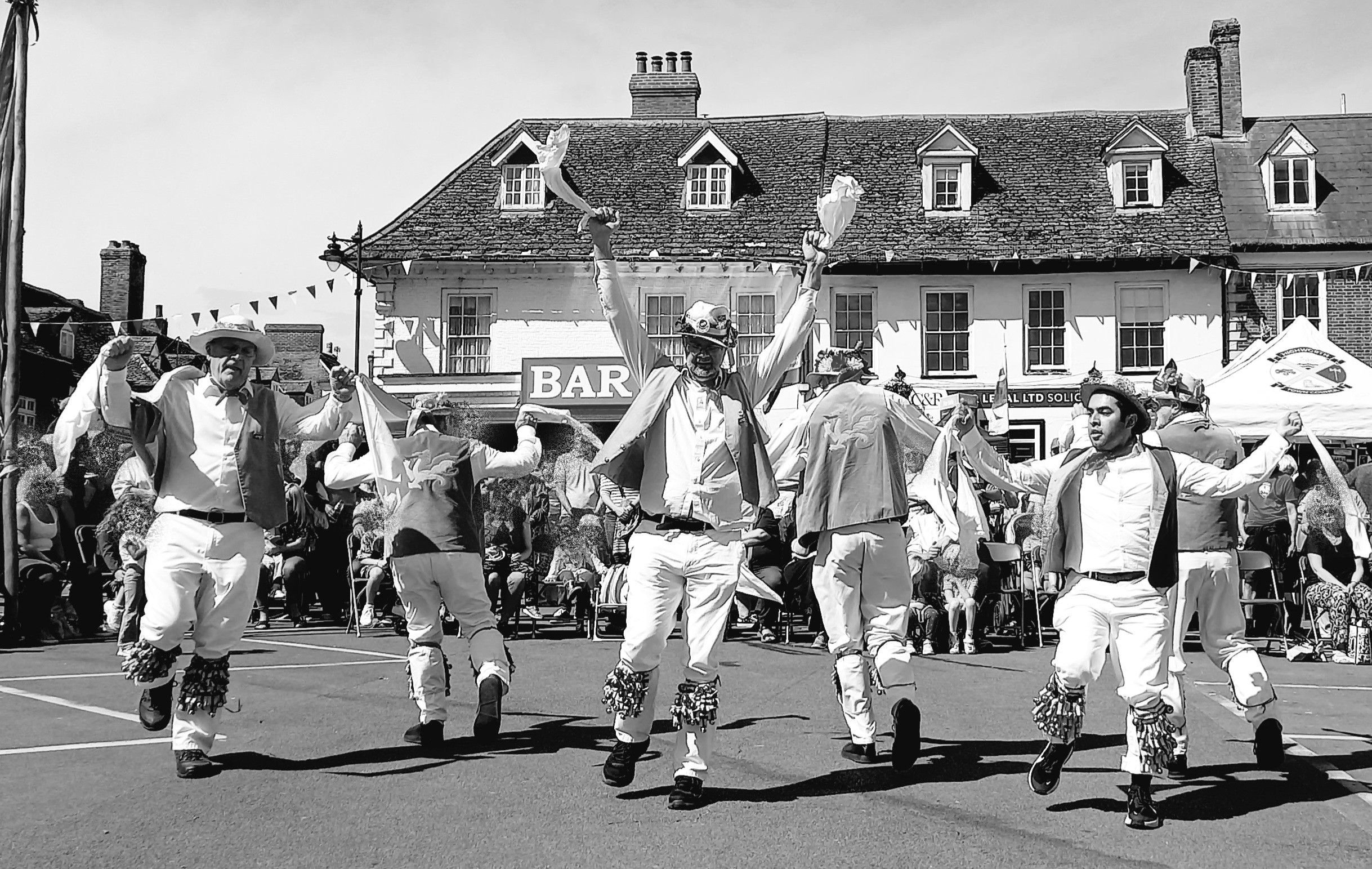 Six bellsteppers waving rags before a crowd overlooked by old shopfronts.