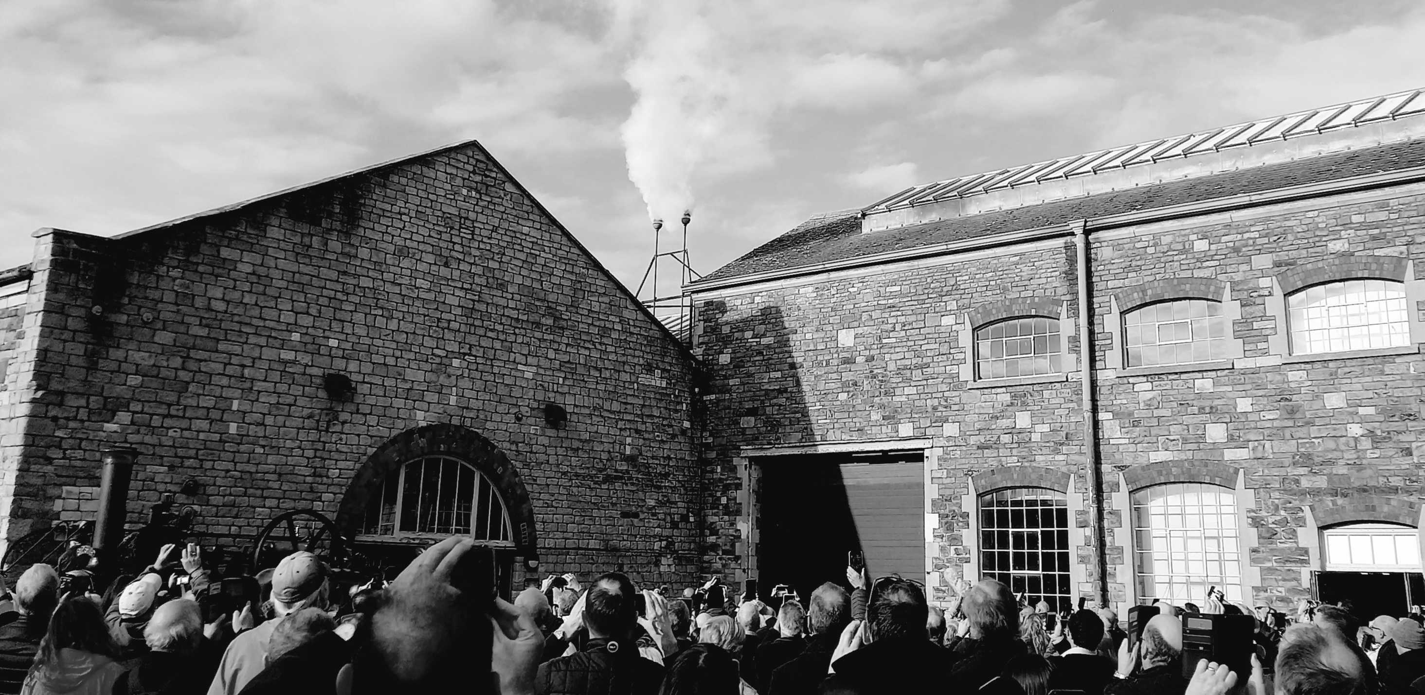 Steam rising from the hooter on the roof of an old brick built building. The backs of heads in the crowd can barely be seen at the bottom.